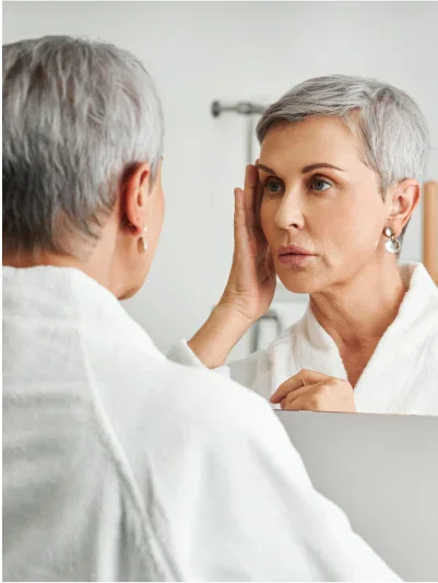 A woman with short, gray hair is looking at her reflection in a mirror. She is wearing a white bathrobe. She is touching her face, and her expression appears somewhat concerned or pensive. The lighting in the bathroom is bright and neutral. The background is a light gray/white bathroom wall. The reflection in the mirror shows a similar-aged woman with a similar expression.
