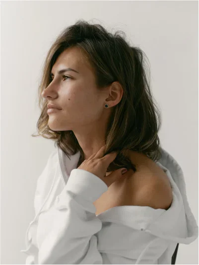 A young woman with brown, shoulder-length hair looks to the left, with her profile visible. She is wearing a white hoodie or top that is pulled down to expose her shoulder, and her hand is resting near her neck, gently touching her hair. She has a dark stud earring in her ear. The background is a plain, light color.