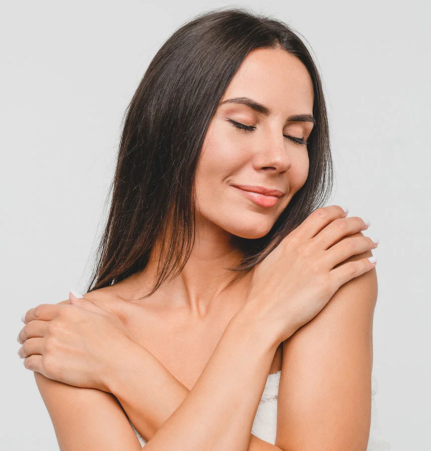 A beautiful woman with long, dark hair gently holds her arms across her chest, with her hands resting on her shoulders. Her eyes are closed, and she has a serene, soft smile on her face, suggesting contentment. She appears to be bare-shouldered, and the background is a plain, light color. - Arm Lift in Watkinsville, GA