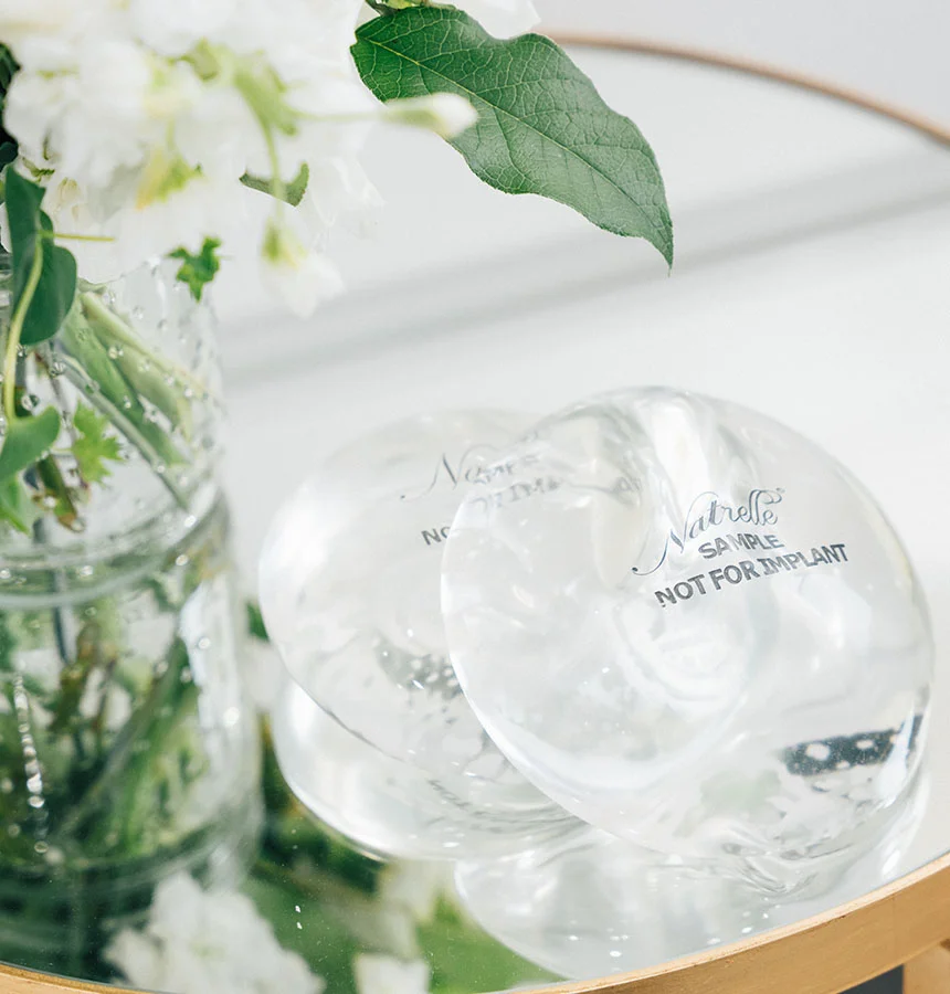 A close-up shot shows two clear, round breast implant samples resting on a reflective surface, likely a mirrored table. The implants have text visible, with "SAMPLE NOT FOR IMPLANT" clearly legible on the front one, and "Natrelle" visible on both. To the left, a vase holding white flowers and green foliage is partially visible. - Breast Implant Revision in Watkinsville, GA
