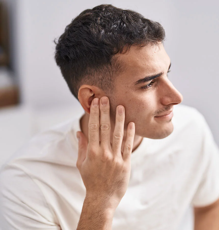 A young man with short, dark curly hair and a light stubble beard is shown in a close-up, looking to his right and smiling slightly. He is wearing a white t-shirt, and his right hand is raised, with fingers spread, touching his earlobe and the side of his face. The background is a soft, light blur. - Earlobe Repair in Watkinsville, GA