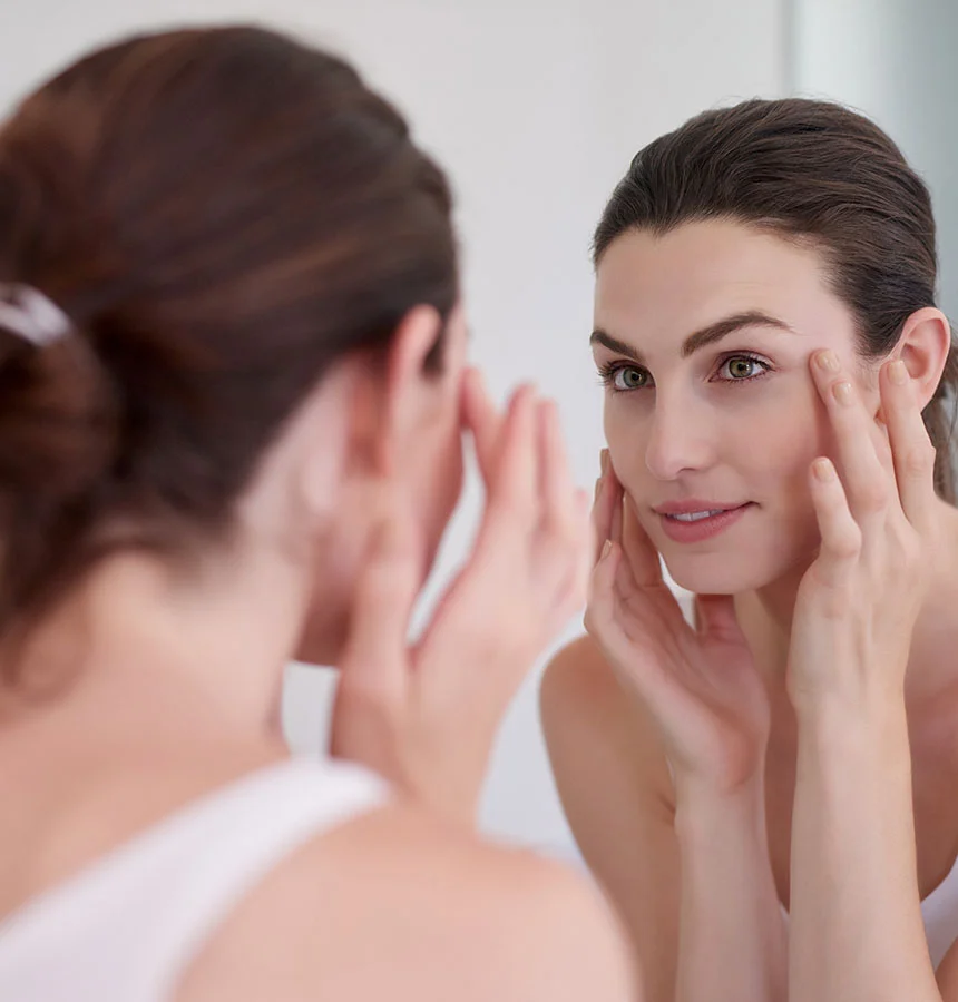 A woman with dark hair pulled back looks at her reflection in a mirror, touching the skin around her eyes with both hands. She is wearing a white tank top. - Eyelid Surgery in Watkinsville, GA