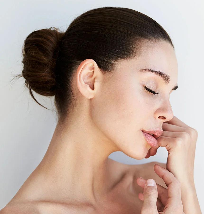 A close up profile shot of a woman with dark hair pulled back in a bun, her eyes closed and head tilted slightly downward. She is bare-shouldered, and her left hand is gently resting on her right shoulder. The background is a plain, light color. - Facial Fat Transfer in Watkinsville, GA