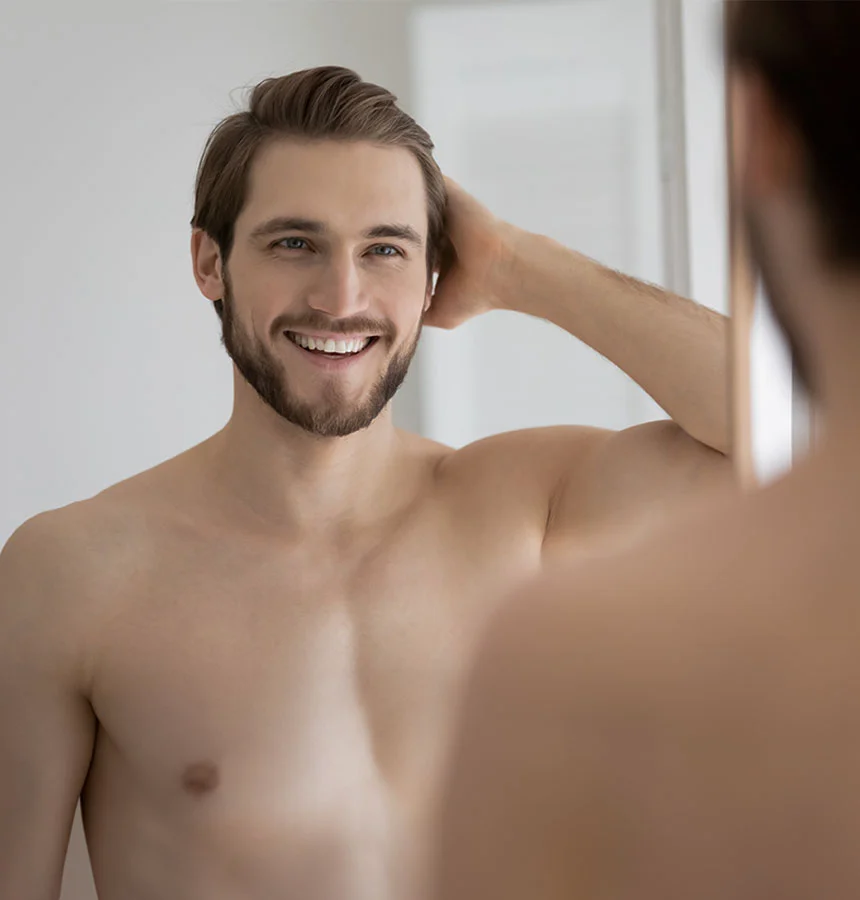 A close-up image shows a young, handsome man with a neatly trimmed beard and well-groomed light brown hair, standing shirtless and smiling broadly while looking at his reflection in a mirror. He has a pleasant expression and one of his hands is raised, running through his hair, suggesting he might be checking his appearance or adjusting his hairstyle. The reflection of his back is visible in the mirror. - Gynecomastia in Watkinsville, GA