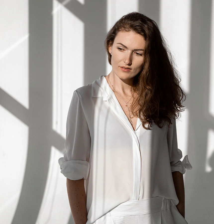 A pensive woman with long, wavy dark hair, wearing a white button-down shirt and white pants, stands against a white wall. Strong sunlight casts distinct geometric shadows on the wall behind her and partially on her, creating a striking visual contrast. She is looking down and to her right, with a thoughtful expression. - Hormone Pellet Therapy in Watkinsville, GA