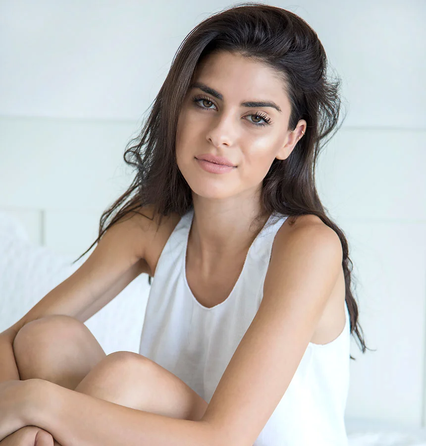 A beautiful young woman with long, dark, slightly wavy hair and piercing eyes looks directly at the camera with a calm expression. She is wearing a simple white tank top and is posed sitting with her knees drawn up. The background is a soft, light, out-of-focus indoor setting, possibly a bed with white headboard and pillows. - Lip Fillers in Watkinsville, GA