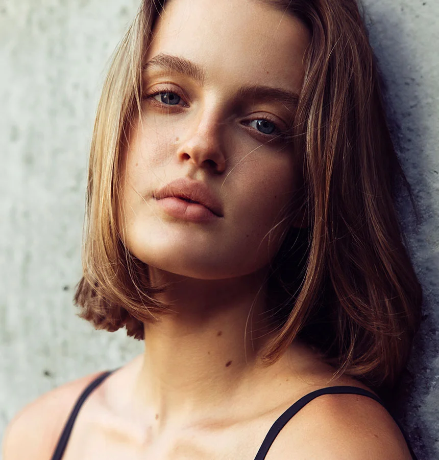 A close-up portrait of a young woman with shoulder-length, light brown hair and blue eyes, looking directly at the viewer with a neutral expression. She has light freckles and is wearing a black camisole. The background appears to be a textured, light gray wall. - Lip Lift in Watkinsville, GA