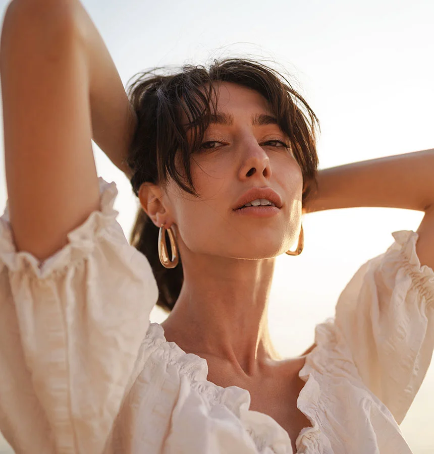 A woman with shoulder-length, dark brown hair is seen from the mid-chest up. She is wearing a off-white, loose-fitting blouse with puffed sleeves. Her hands are resting on her head, and her expression is neutral, though slightly directed toward the viewer. The lighting suggests a golden hour, with a soft, warm glow. She is wearing gold hoop earrings. Her skin appears smooth and light-toned. - Neck Lift in Watkinsville, GA