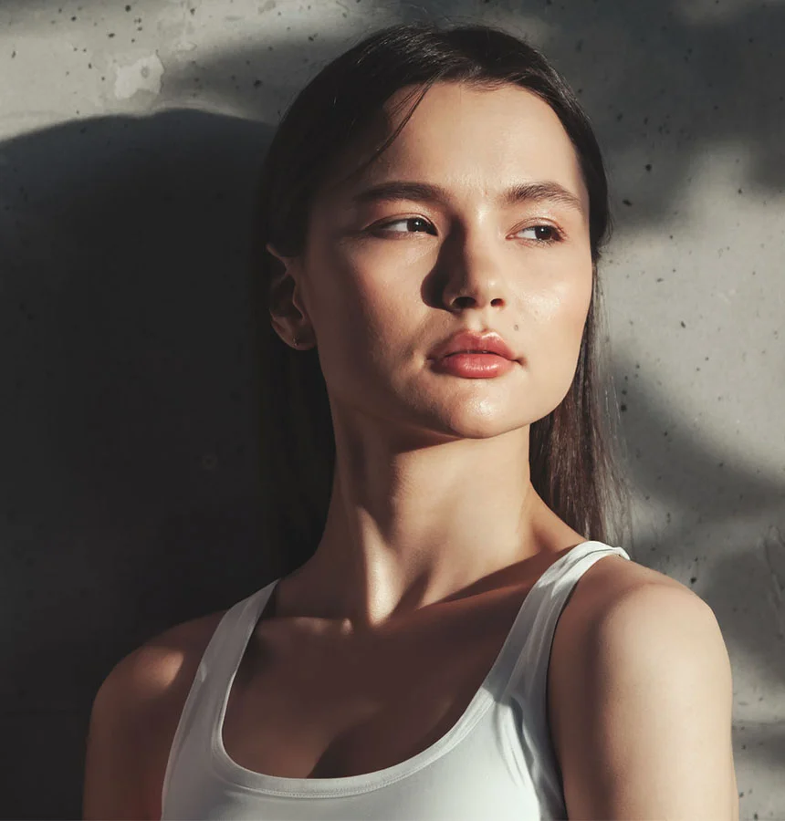 A woman with dark brown hair is seen from the mid-chest up against a textured light gray concrete wall. Her head is turned slightly to the right as she gazes into the distance. Dramatic lighting casts strong highlights on her face and hair, emphasizing her smooth, light-toned skin and subtle, natural makeup with rosy-pink lips. She appears bare-shouldered. - Skin Resurfacing in Watkinsville, GA